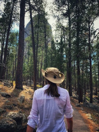 Hiker seen from behind on a pine forest trail in a tan wide-brim hat and a white long-sleeve shirt