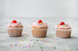 Three cupcakes with pink frosting and colorful sprinkles, topped with a red cherry, are arranged in a line on a light-colored surface sprinkled with confetti.