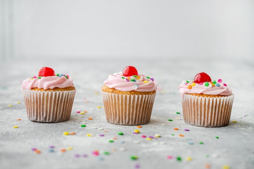 Three cupcakes with pink frosting and colorful sprinkles, topped with a red cherry, are arranged in a line on a light-colored surface sprinkled with confetti.