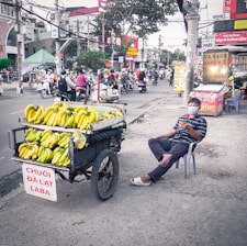 A street vendor sits on a plastic chair beside a cart loaded with bunches of bananas on a bustling city street. The vendor wears a striped shirt and a face mask, holding a smartphone. The scene is in an urban setting with various businesses and people on motorcycles in the background.