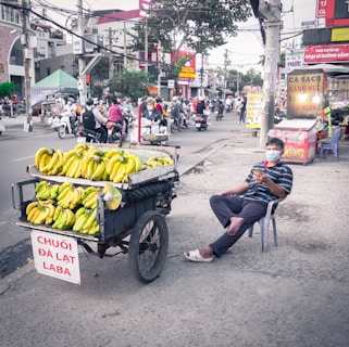 A street vendor sits on a plastic chair beside a cart loaded with bunches of bananas on a bustling city street. The vendor wears a striped shirt and a face mask, holding a smartphone. The scene is in an urban setting with various businesses and people on motorcycles in the background.