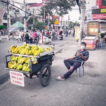 A street vendor sits on a plastic chair beside a cart loaded with bunches of bananas on a bustling city street. The vendor wears a striped shirt and a face mask, holding a smartphone. The scene is in an urban setting with various businesses and people on motorcycles in the background.