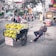 A street vendor sits on a plastic chair beside a cart loaded with bunches of bananas on a bustling city street. The vendor wears a striped shirt and a face mask, holding a smartphone. The scene is in an urban setting with various businesses and people on motorcycles in the background.