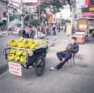 A street vendor sits on a plastic chair beside a cart loaded with bunches of bananas on a bustling city street. The vendor wears a striped shirt and a face mask, holding a smartphone. The scene is in an urban setting with various businesses and people on motorcycles in the background.
