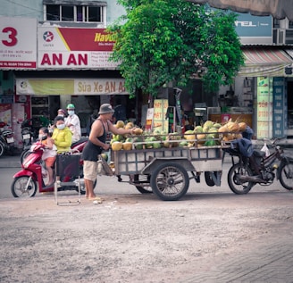 A street scene in an urban environment featuring a man selling coconuts from a cart attached to a motorcycle. A family with children is seated on a red scooter nearby, and several signs with Vietnamese text are visible in the background. A small tree provides some greenery amidst the bustling atmosphere.