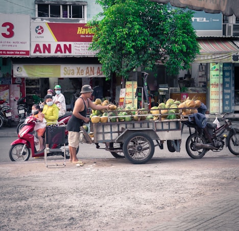 A street scene in an urban environment featuring a man selling coconuts from a cart attached to a motorcycle. A family with children is seated on a red scooter nearby, and several signs with Vietnamese text are visible in the background. A small tree provides some greenery amidst the bustling atmosphere.