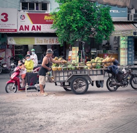 A street scene in an urban environment featuring a man selling coconuts from a cart attached to a motorcycle. A family with children is seated on a red scooter nearby, and several signs with Vietnamese text are visible in the background. A small tree provides some greenery amidst the bustling atmosphere.