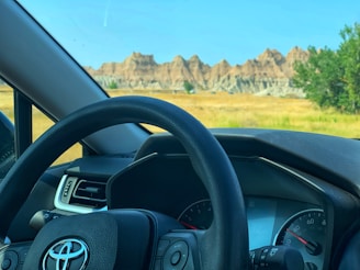 A car dashboard and steering wheel with a Toyota logo are visible from inside the vehicle. Outside, there is a scenic view of rocky formations and a grassy field under a clear blue sky.