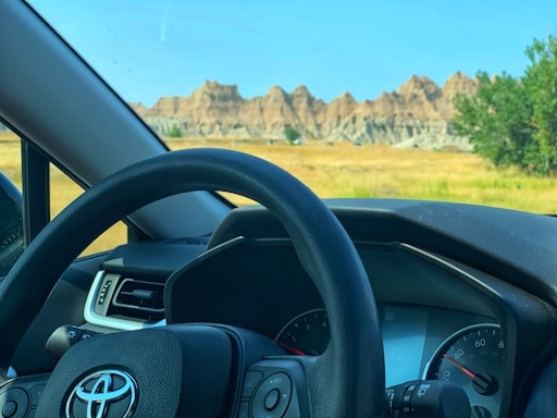 A car dashboard and steering wheel with a Toyota logo are visible from inside the vehicle. Outside, there is a scenic view of rocky formations and a grassy field under a clear blue sky.