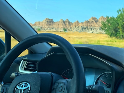 A car dashboard and steering wheel with a Toyota logo are visible from inside the vehicle. Outside, there is a scenic view of rocky formations and a grassy field under a clear blue sky.