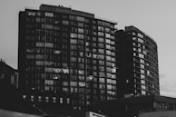 A black and white photograph captures two tall, modern apartment buildings with numerous windows. Some windows are illuminated, indicating lights on inside the apartments. The structures have a sleek, urban design and dominate the skyline.
