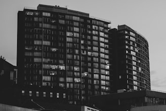 A black and white photograph captures two tall, modern apartment buildings with numerous windows. Some windows are illuminated, indicating lights on inside the apartments. The structures have a sleek, urban design and dominate the skyline.
