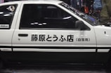 Side view of a classic white car featuring Japanese text on its door. The car has a two-tone design with black accents, and there is a sticker on the rear window. The background is slightly blurred with people and a dimly lit environment.