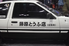 Side view of a classic white car featuring Japanese text on its door. The car has a two-tone design with black accents, and there is a sticker on the rear window. The background is slightly blurred with people and a dimly lit environment.