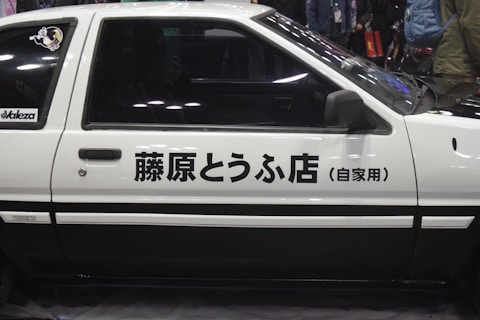 Side view of a classic white car featuring Japanese text on its door. The car has a two-tone design with black accents, and there is a sticker on the rear window. The background is slightly blurred with people and a dimly lit environment.