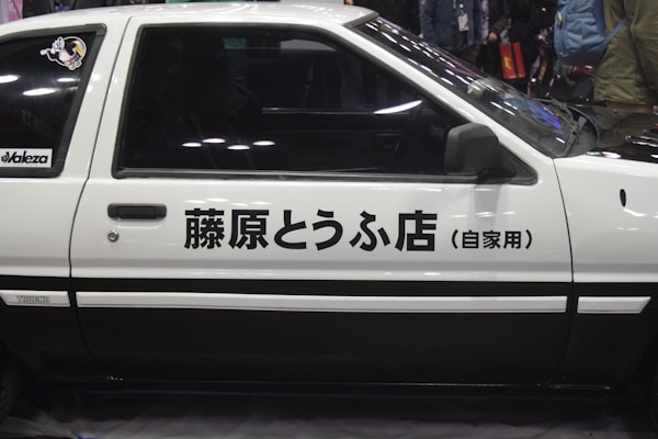 Side view of a classic white car featuring Japanese text on its door. The car has a two-tone design with black accents, and there is a sticker on the rear window. The background is slightly blurred with people and a dimly lit environment.