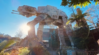 A large, mechanical, four-legged walker stands among trees and foliage under a clear blue sky. Sunlight filters through the trees, casting highlights on the metallic structure, surrounded by a wooden fence and bushes.