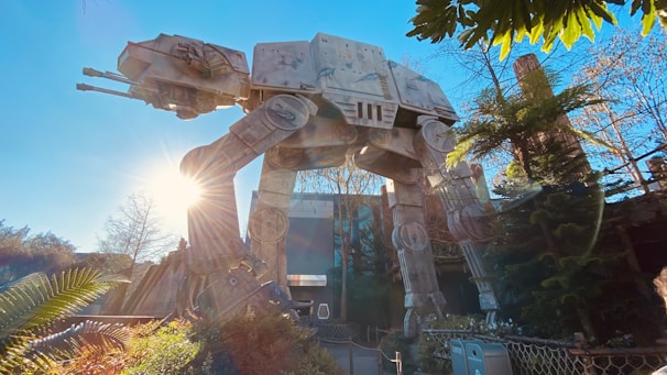 A large, mechanical, four-legged walker stands among trees and foliage under a clear blue sky. Sunlight filters through the trees, casting highlights on the metallic structure, surrounded by a wooden fence and bushes.