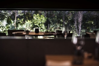 A dimly lit restaurant interior with a round table set for dining. The table is adorned with glasses and a small centerpiece. Large windows provide a view of lush greenery outside, creating a serene atmosphere.