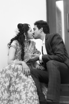 A couple is seated on stairs, leaning towards each other for a kiss. The woman is wearing a traditional embroidered dress with jewelry, including earrings and a hair accessory. The man is in a formal suit.