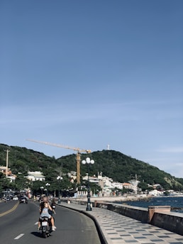 A coastal street scene featuring a motorcyclist riding on a road beside the ocean. There is a large hill covered in greenery in the background, with a crane and a statue atop the hill. Streetlights line the road, and various buildings can be seen along the coastline.