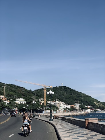 A coastal street scene featuring a motorcyclist riding on a road beside the ocean. There is a large hill covered in greenery in the background, with a crane and a statue atop the hill. Streetlights line the road, and various buildings can be seen along the coastline.