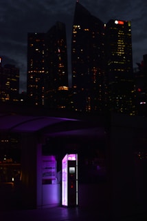 An illuminated kiosk with pink and purple lighting is situated outdoors at night with tall, dark skyscrapers in the background. The buildings are dotted with numerous small, lit-up windows.