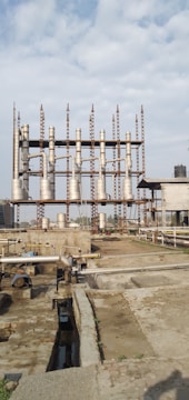 An industrial site with multiple tall, cylindrical metal structures and piping systems. The structures are elevated on a framework of girders. The ground appears to be a concrete platform with additional piping and ducts. The sky above is partly cloudy.