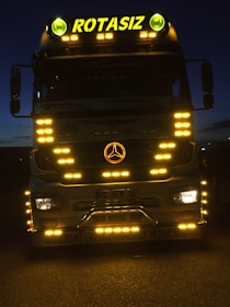 A large truck with multiple bright yellow lights around its front. The word 'ROTASIZ' is illuminated on top of the truck. Some details and symbols, including a central emblem, are visible on the grill and bumper. The vehicle is stationary against a dark background, giving a sense of night or low-light conditions.
