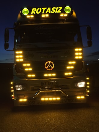 A large truck with multiple bright yellow lights around its front. The word 'ROTASIZ' is illuminated on top of the truck. Some details and symbols, including a central emblem, are visible on the grill and bumper. The vehicle is stationary against a dark background, giving a sense of night or low-light conditions.