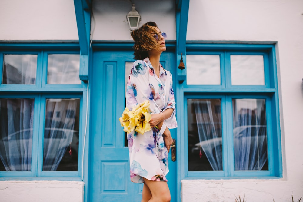 woman in white and blue floral dress holding yellow flower bouquet standing beside blue wooden window