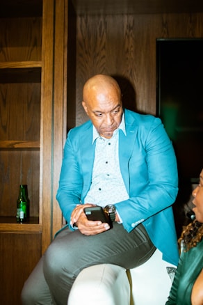 A man in a blue blazer and light blue shirt is sitting with his legs crossed, holding a smartphone. He is looking intently at the device. The background includes wooden shelves and a green bottle, likely a beverage. Another person is partially visible, engaged in conversation.