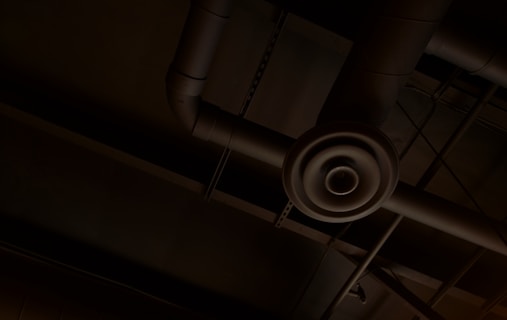 An industrial ceiling with exposed ductwork and a circular air vent. The pipes and ducts create a network of lines and curves against the dark background.