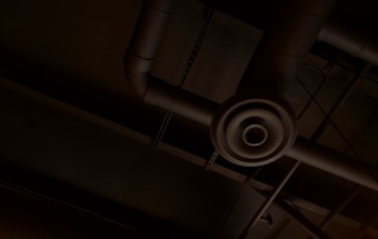 An industrial ceiling with exposed ductwork and a circular air vent. The pipes and ducts create a network of lines and curves against the dark background.