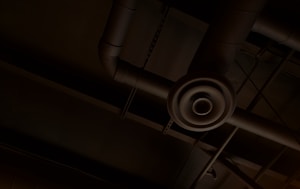 An industrial ceiling with exposed ductwork and a circular air vent. The pipes and ducts create a network of lines and curves against the dark background.