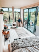 A cozy indoor space with a modern, rustic design featuring large glass doors opening to a lush green field. A small wood-burning stove is on one side, next to a comfortable armchair with a dark pillow. The floor is adorned with a patterned rug, and plants add a touch of greenery. Decorative items and books are arranged on a wooden side table.