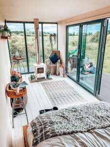A cozy indoor space with a modern, rustic design featuring large glass doors opening to a lush green field. A small wood-burning stove is on one side, next to a comfortable armchair with a dark pillow. The floor is adorned with a patterned rug, and plants add a touch of greenery. Decorative items and books are arranged on a wooden side table.