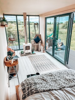 A cozy indoor space with a modern, rustic design featuring large glass doors opening to a lush green field. A small wood-burning stove is on one side, next to a comfortable armchair with a dark pillow. The floor is adorned with a patterned rug, and plants add a touch of greenery. Decorative items and books are arranged on a wooden side table.
