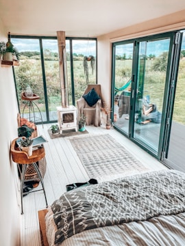 A cozy indoor space with a modern, rustic design featuring large glass doors opening to a lush green field. A small wood-burning stove is on one side, next to a comfortable armchair with a dark pillow. The floor is adorned with a patterned rug, and plants add a touch of greenery. Decorative items and books are arranged on a wooden side table.