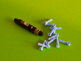 A black and beige lancing device is placed on a bright green surface alongside multiple blue and white lancets scattered around.