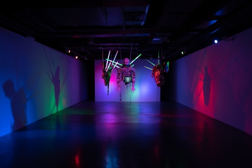 An art installation in a dimly lit room features abstract sculptures suspended from the ceiling, with neon lights casting colorful shadows on the walls. The scene is illuminated by vibrant hues of green, blue, pink, and red, creating a surreal and immersive atmosphere.
