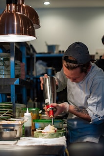 A chef in a kitchen is carefully using a cream dispenser over a dish. The setting is a bustling kitchen with various cooking tools, ingredients, and dishes arranged nearby.