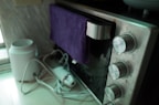 An oven with large silver knobs is partially covered with a purple towel hanging over its handle. There is a white appliance with attached cables on the left, possibly an electric kettle or air purifier, resting on a countertop.