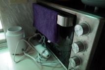 An oven with large silver knobs is partially covered with a purple towel hanging over its handle. There is a white appliance with attached cables on the left, possibly an electric kettle or air purifier, resting on a countertop.