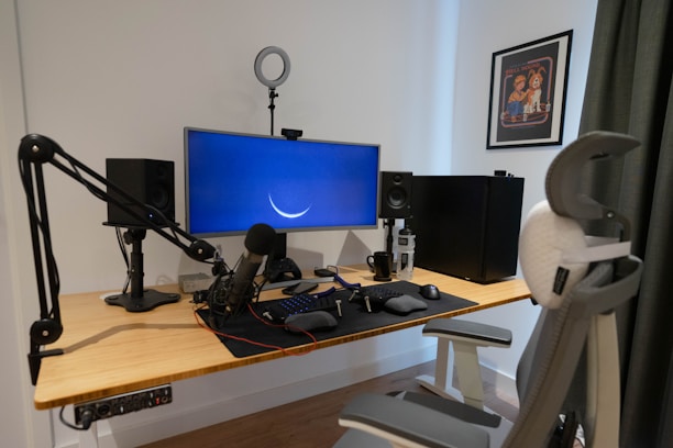 A modern workspace setup featuring a wooden desk with a mounted microphone on an adjustable arm, dual speakers, a large monitor displaying a crescent shape, and a keyboard and mouse. A large ergonomic office chair faces the desk, which includes multiple tech gadgets and accessories. Wall art and a curtain are visible in the background.