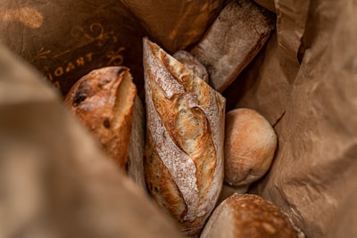 A variety of freshly baked bread inside a brown paper bag. The assortment includes a prominently visible crusty baguette with a golden-brown crust and flour dusting. Additional loaves of different bread types, including a round bun, are also present, all exhibiting a rustic and artisanal appearance.