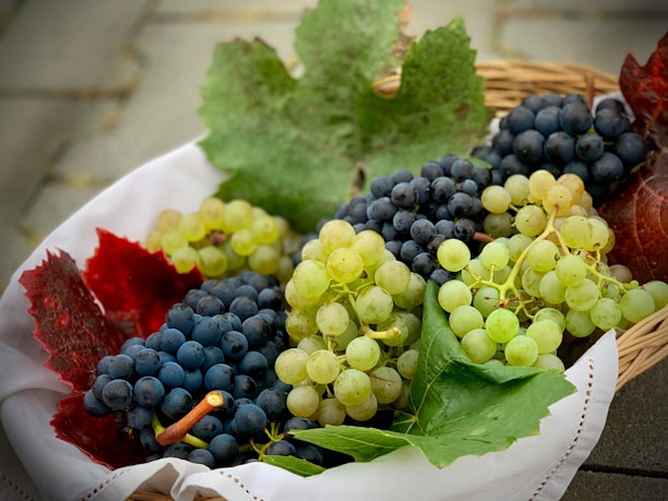 A wicker basket contains clusters of dark and light grapes, surrounded by large green and red grape leaves. The basket is lined with a white cloth, and the rustic setting suggests a harvest or autumn theme.