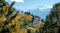 A large, elegant villa is situated in a picturesque mountain landscape. Surrounded by vibrant green trees and a winding pathway leading to the building, the scene features majestic snow-capped mountains in the background under a clear blue sky. The lush greenery and serene environment suggest a peaceful and secluded location.