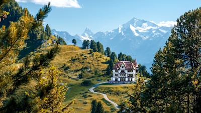 A large, elegant villa is situated in a picturesque mountain landscape. Surrounded by vibrant green trees and a winding pathway leading to the building, the scene features majestic snow-capped mountains in the background under a clear blue sky. The lush greenery and serene environment suggest a peaceful and secluded location.