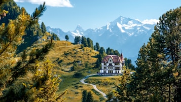 A large, elegant villa is situated in a picturesque mountain landscape. Surrounded by vibrant green trees and a winding pathway leading to the building, the scene features majestic snow-capped mountains in the background under a clear blue sky. The lush greenery and serene environment suggest a peaceful and secluded location.